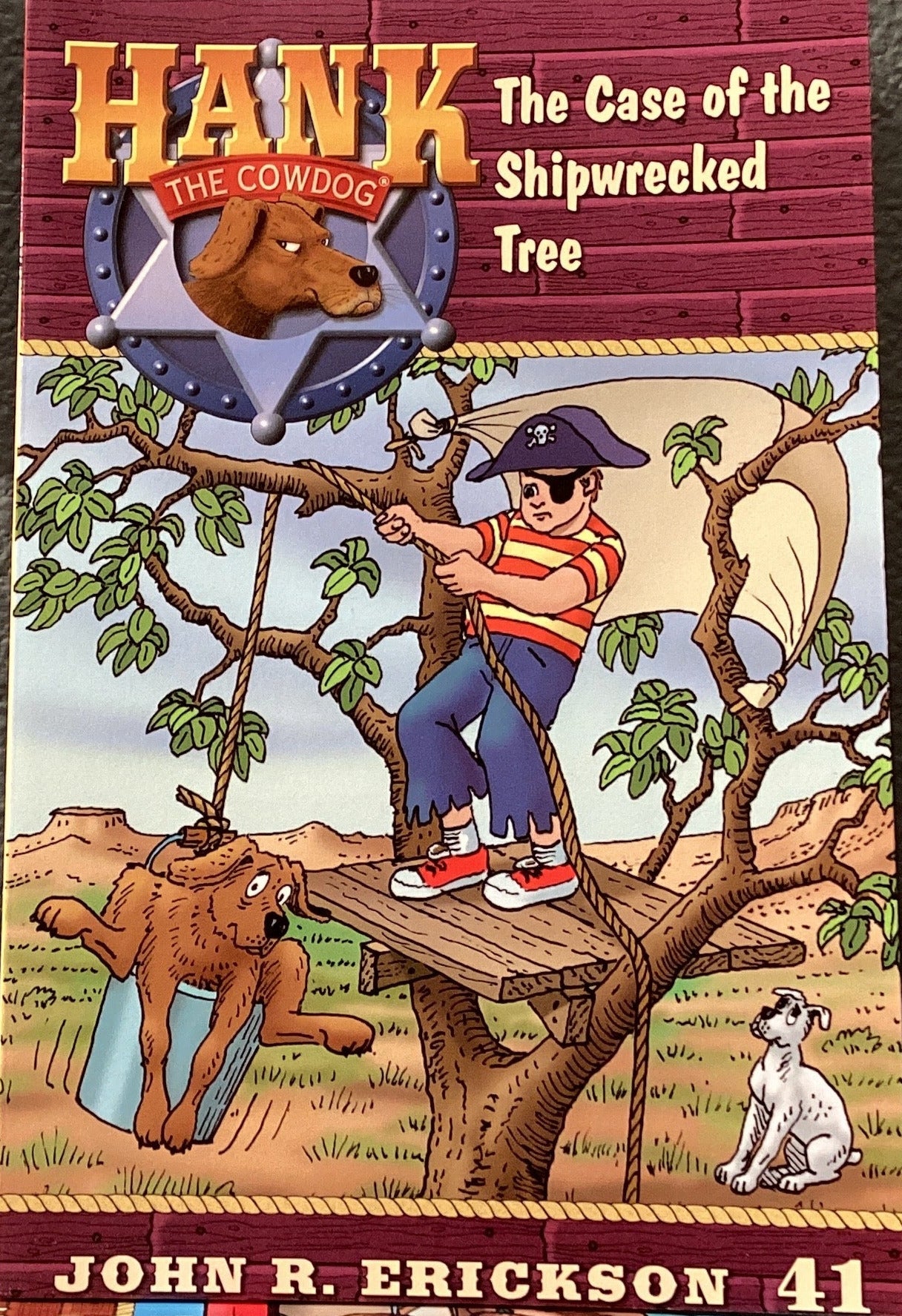 Book titled 'Hank the Cowdog: The Case of the Shipwrecked Tree' by John R. Erickson on a table against a dark wall.