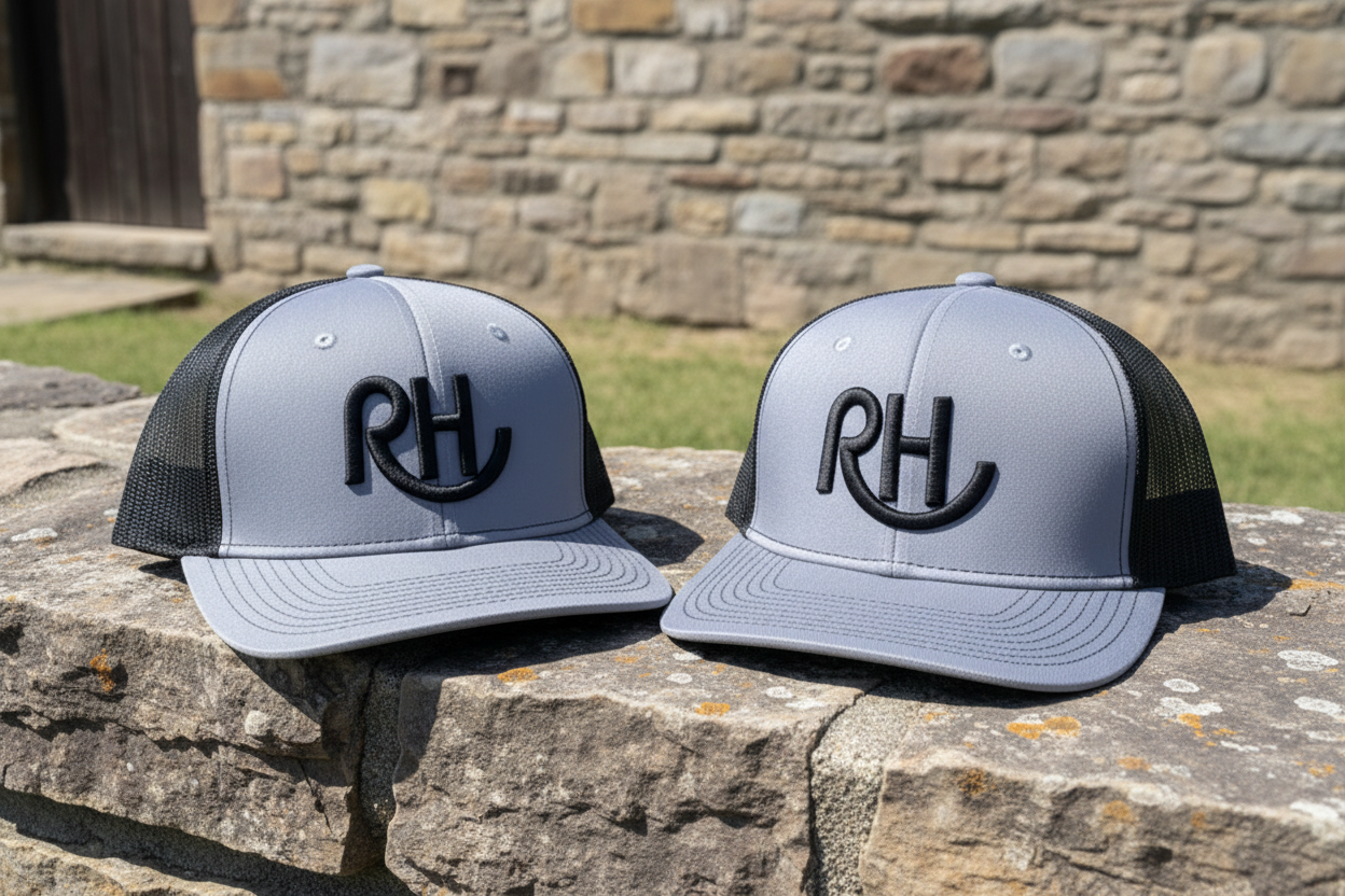 Two gray and black hats with 'RH' logo on a wooden post with a grassy field and cows in the background.