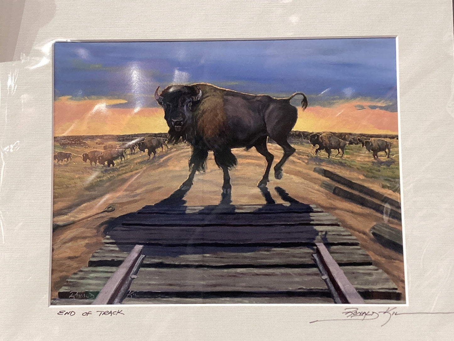 Framed artwork of a bison on train tracks against a sunset landscape.