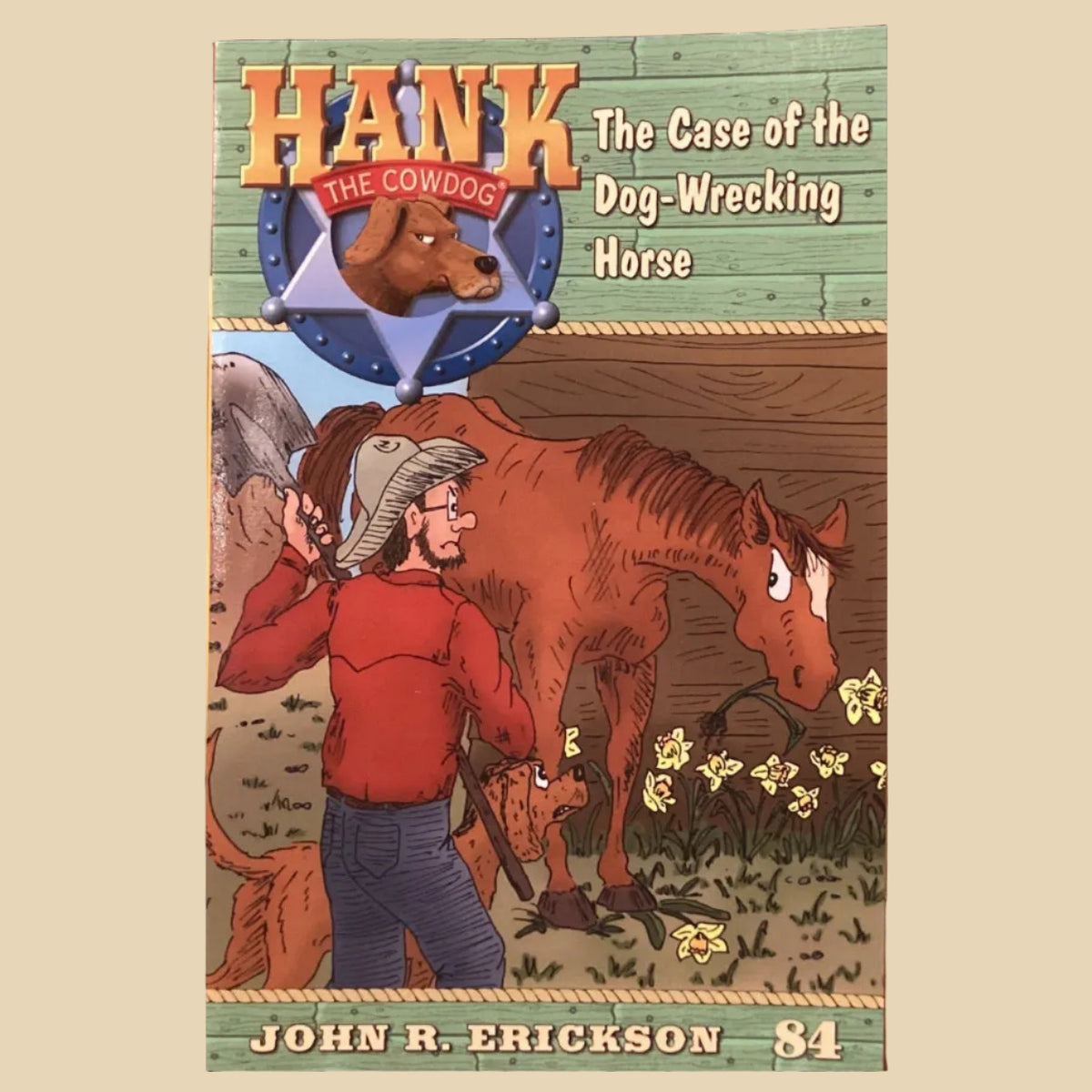 The front cover of the book 'Hank the Cowboy: The Case of the Dog-Wrecking Horse' by John R. Erickson is displayed upright on a small white stand, positioned in the center of a wooden bookshelf lined with copies of the book.