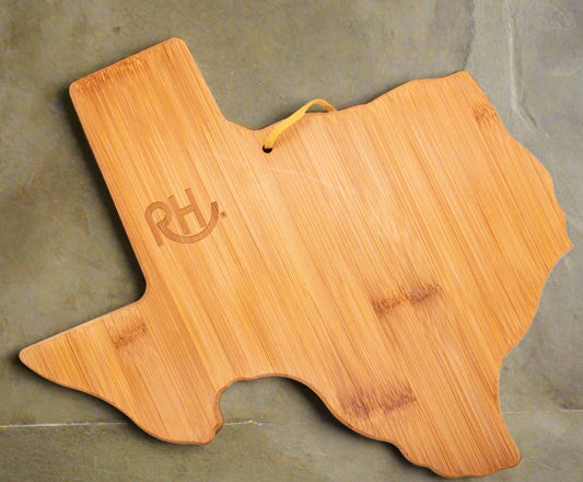 A bamboo cutting board in the shape of the state of Texas, with a branded logo on the top surface and a hanging loop.
