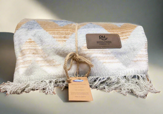A folded blanket with a striped pattern, predominantly beige and white, with a fringe and a brand tag that reads 'RH'.