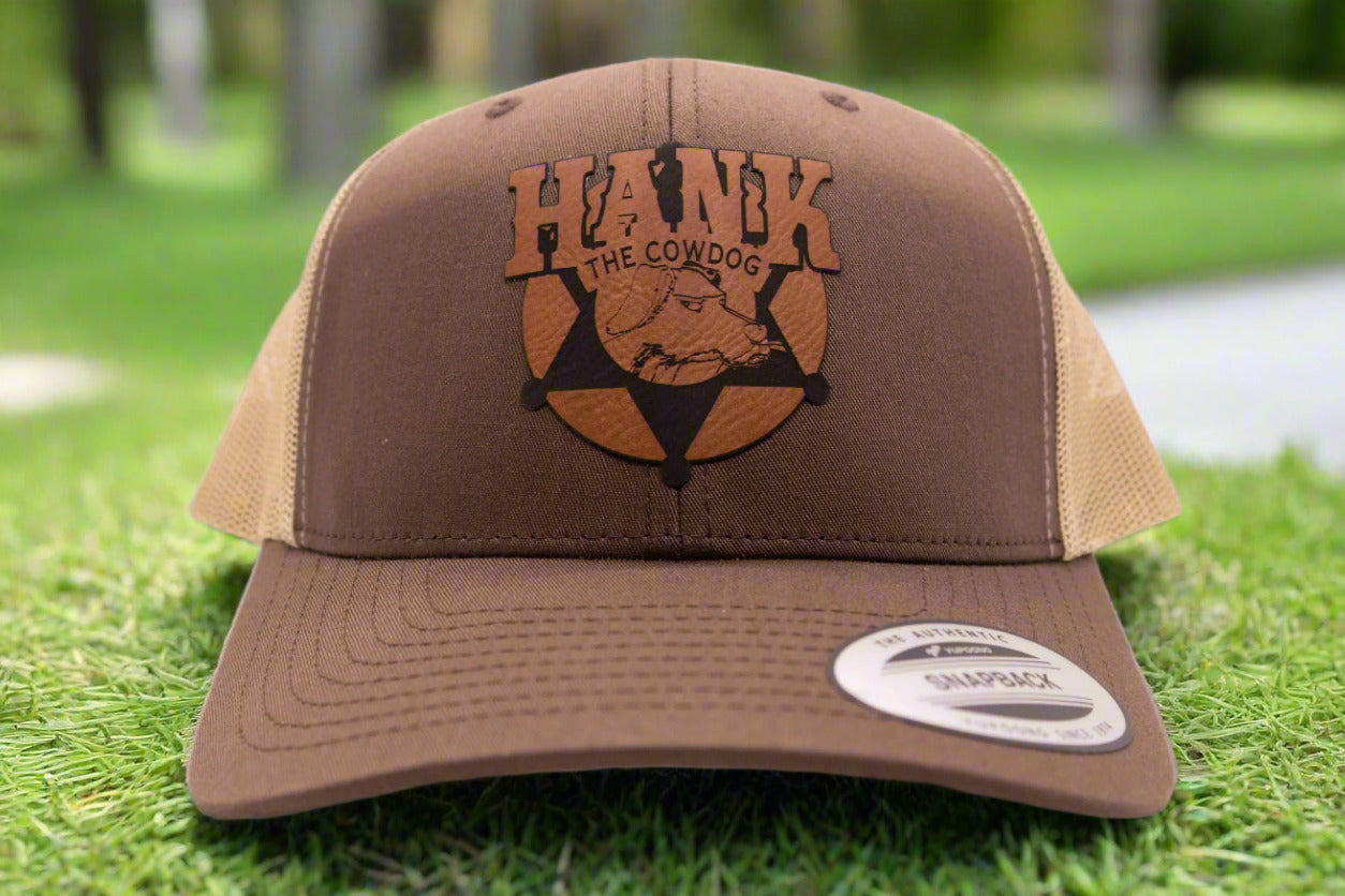 A brown and beige trucker cap with a leather patch featuring text 'HANK THE COWDOG'.