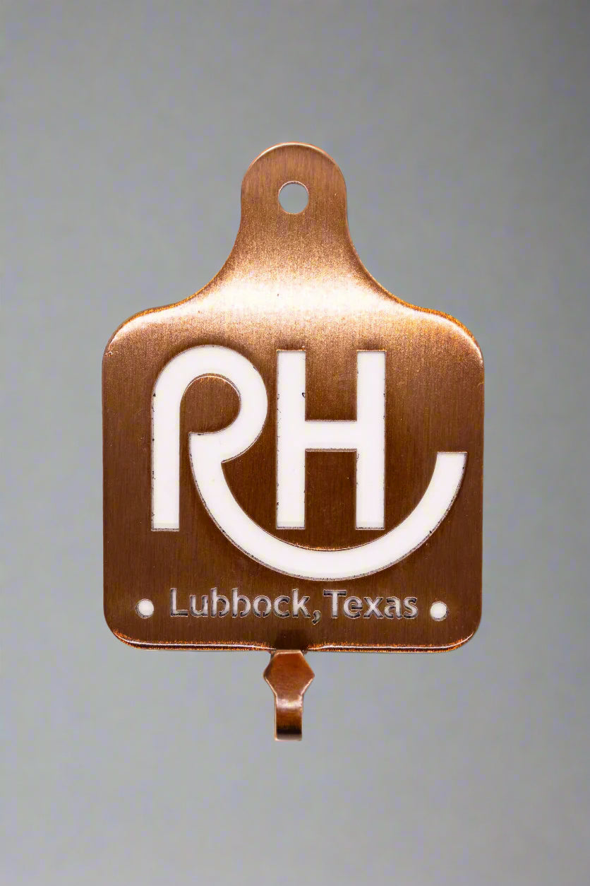 A bronze hook with the letters 'RH' and the text 'Lubbock, Texas' on it, mounted on a wall.