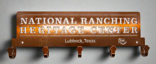 A metal coat rack with five hooks, featuring text that reads 'National Ranching Heritage Center, Lubbock, Texas'. The coat rack has a bronze finish.