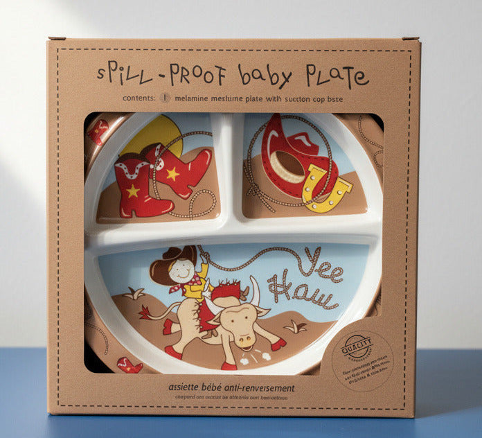 A boxed spill-proof baby plate set displayed on a shelf against a gray wall. The packaging is tan cardboard with a clear window showing a white divided plate decorated with cowboy-themed illustrations, including red boots with yellow stars, a horseshoe, and a light blue lower section with a cowboy and calf and the words 'Vee Hawi'.
