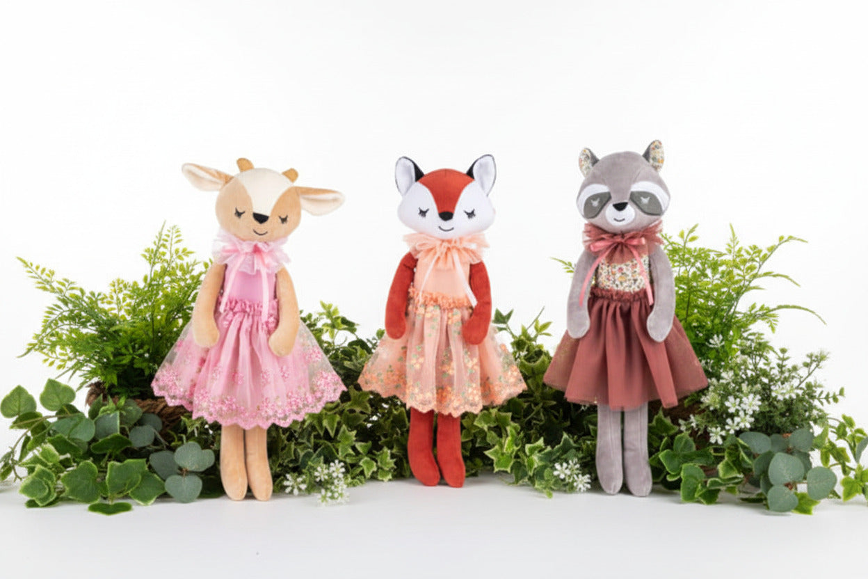 Three plush animal dolls standing side by side against a white background: a tan deer in a pink dress, a red fox in a peach dress, and a gray raccoon in a dusty rose dress.