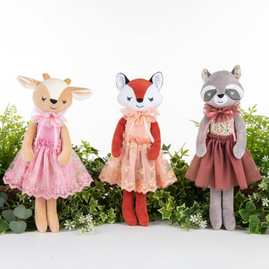Three plush animal dolls standing side by side against a white background: a tan deer in a pink dress, a red fox in a peach dress, and a gray raccoon in a dusty rose dress.