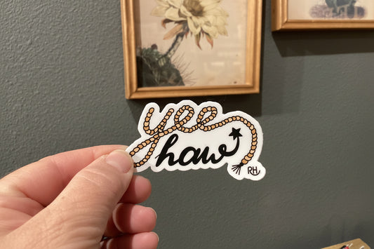 Hand holding a sticker with 'Yee haw' design against a wall with framed pictures.