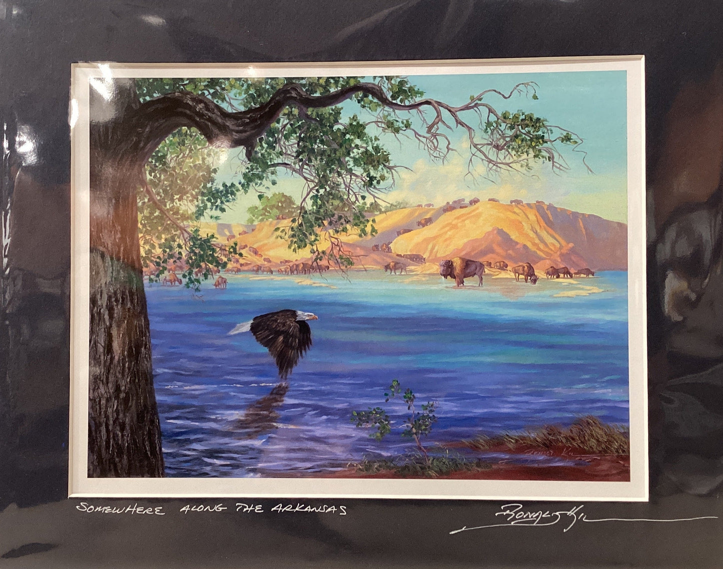 Framed artwork of a landscape with a tree, water, and mountains.