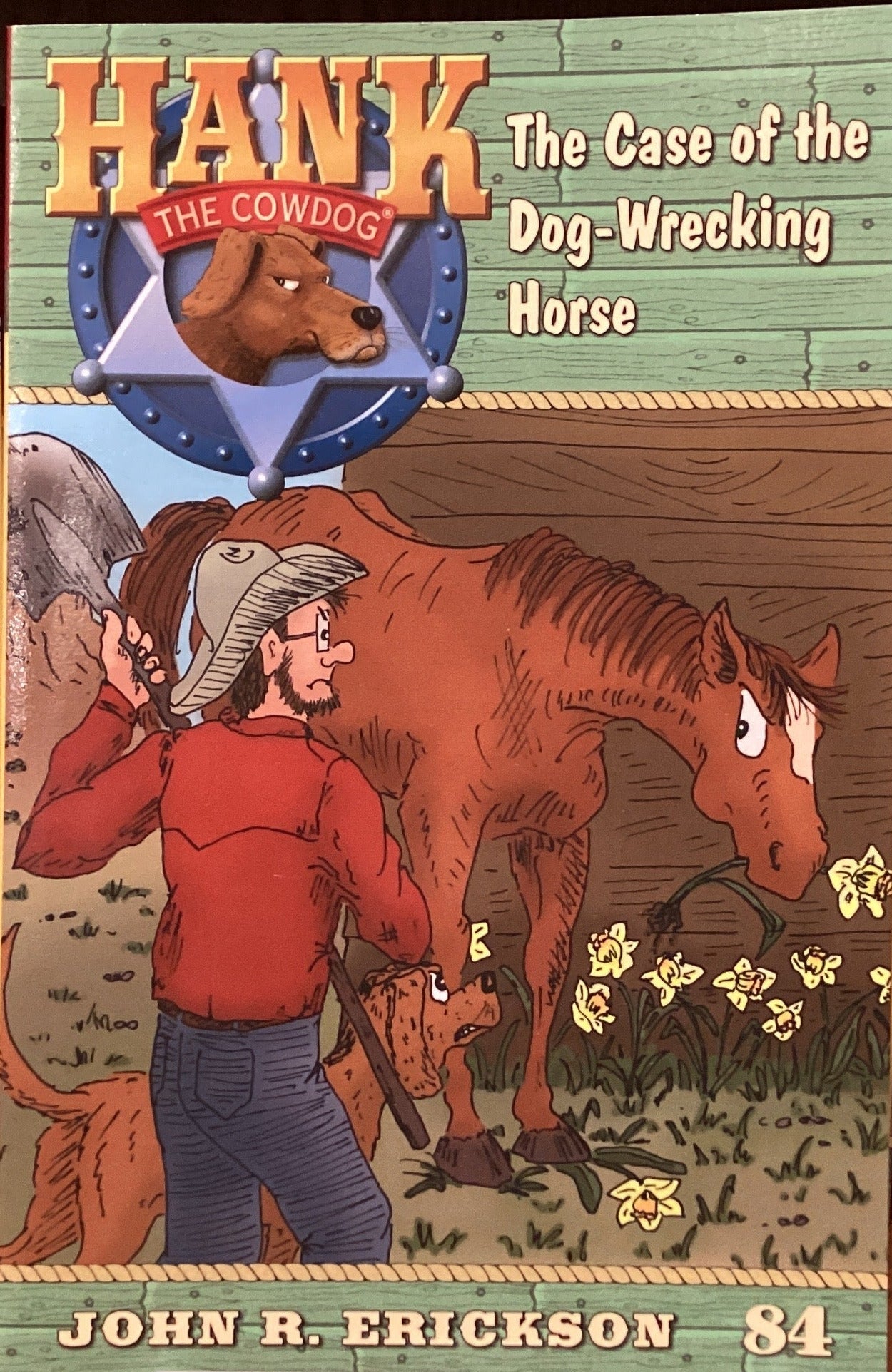 The front cover of the book 'Hank the Cowboy: The Case of the Dog-Wrecking Horse' by John R. Erickson is displayed upright on a small white stand, positioned in the center of a wooden bookshelf lined with copies of the book.