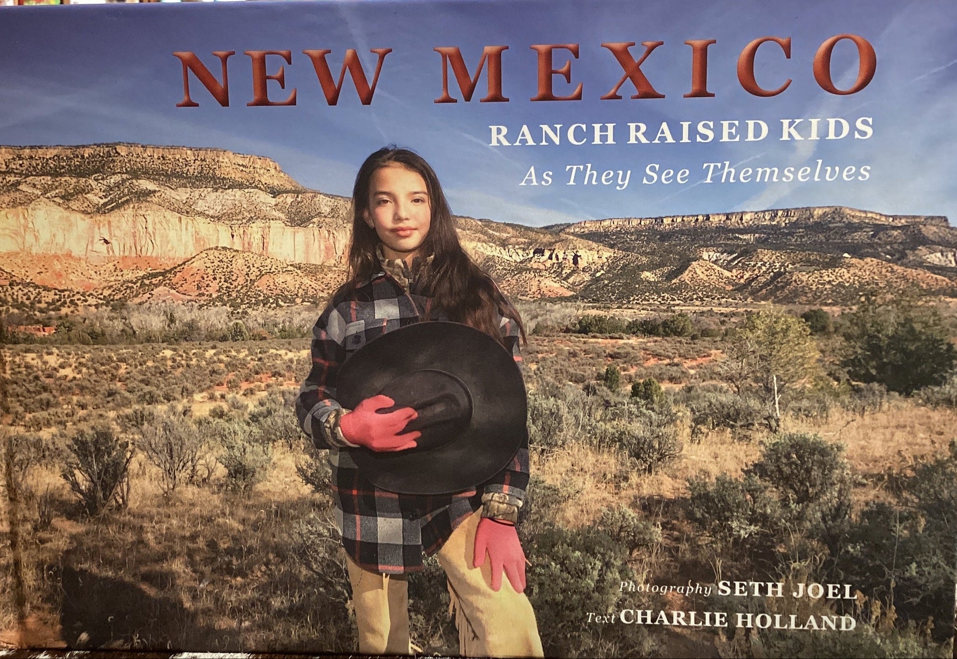 Book titled 'New Mexico Ranch Raised Kids' on a surface with a bookshelf background
