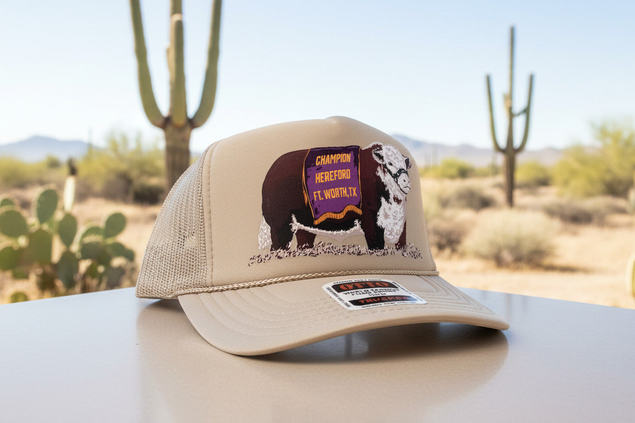 A beige trucker hat with a graphic of a cow on the front panel, wearing a purple banner that reads 'CHAMPION HEREFORD FT. WORTH, TX'.