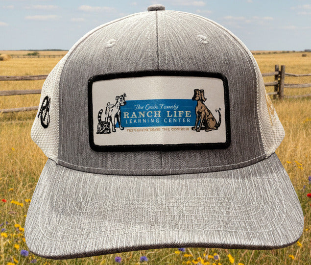 A heather gray trucker hat with a rectangular front patch reading 'The Great Ranch Life Learning Center' and featuring dog illustrations, displayed indoors on a stand.