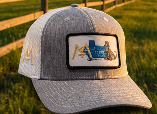 A gray and white trucker cap with a gold embroidered 'M' on the front, displayed in a retail store setting with other apparel items hanging in the background.