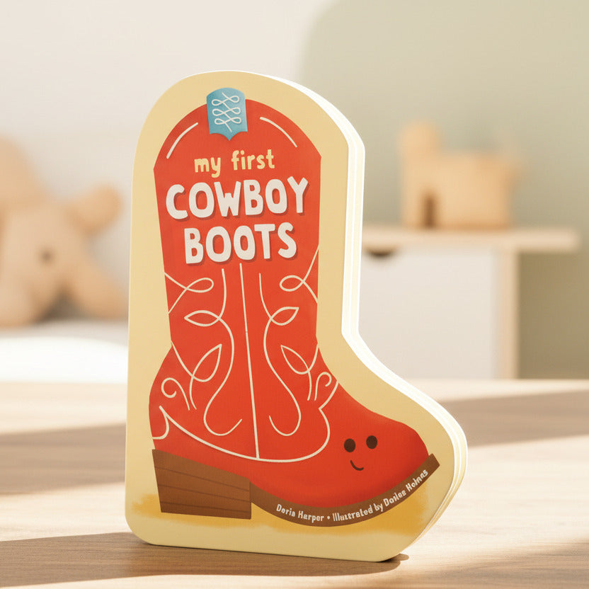 A children's board book shaped like a cowboy boot titled "my first COWBOY BOOTS" by Dorie Norper, illustrated by Danielle Hackett. The book features a bright orange boot design with white decorative stitching, a yellow cover, and a small smiling face on the heel.