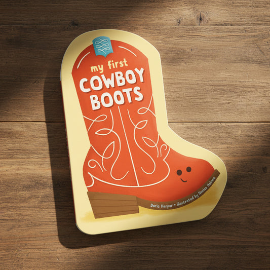 A children's board book shaped like a cowboy boot titled "my first COWBOY BOOTS" by Dorie Norper, illustrated by Danielle Hackett. The book features a bright orange boot design with white decorative stitching, a yellow cover, and a small smiling face on the heel.