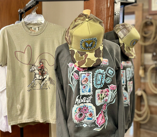 T-shirt with a graphic design of a person on a horse, a cap, and a sweatshirt with colorful designs on a rack.