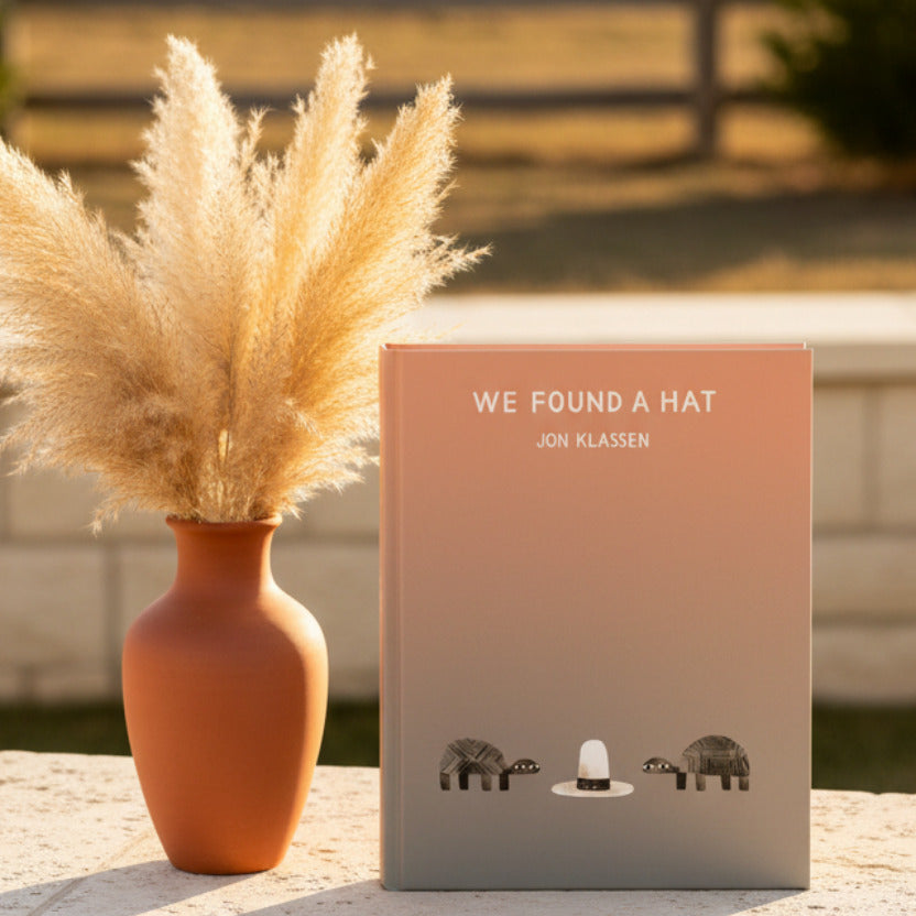 A minimalist art print titled 'WE FOUND A HAT' by Jon Klassen, featuring a peach-to-gray gradient background with a white cowboy hat centered near the bottom, flanked by two small dark patterned animals.